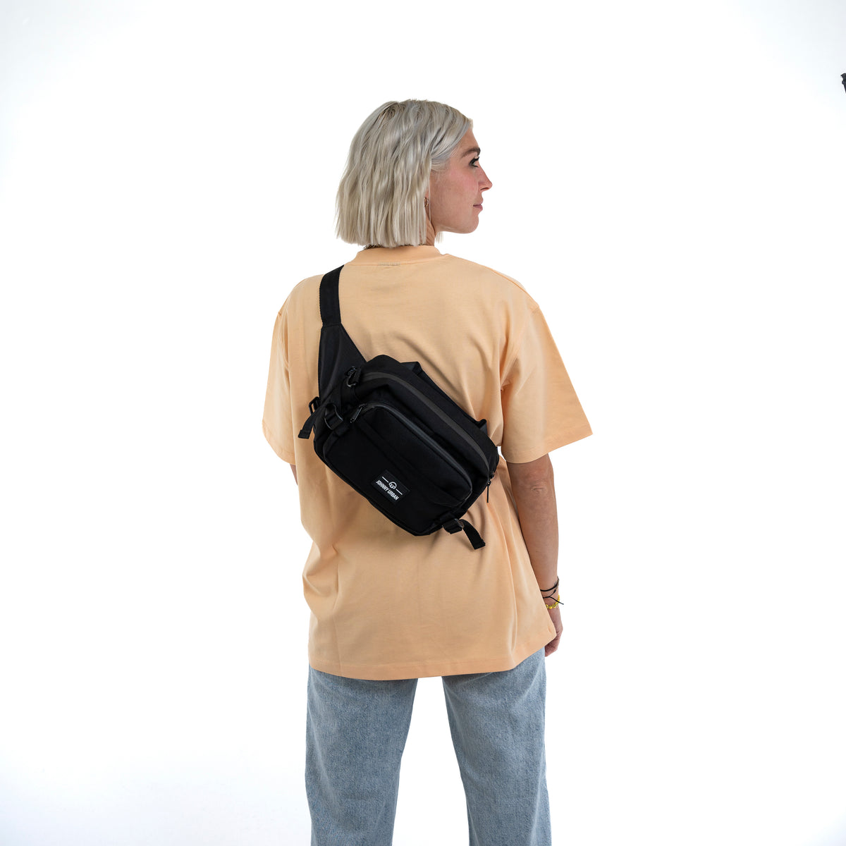 Frau trägt schwarze Johnny Urban Bauchtasche 'Travis' diagonal am Rücken mit beiger T-Shirt und blauen Jeans