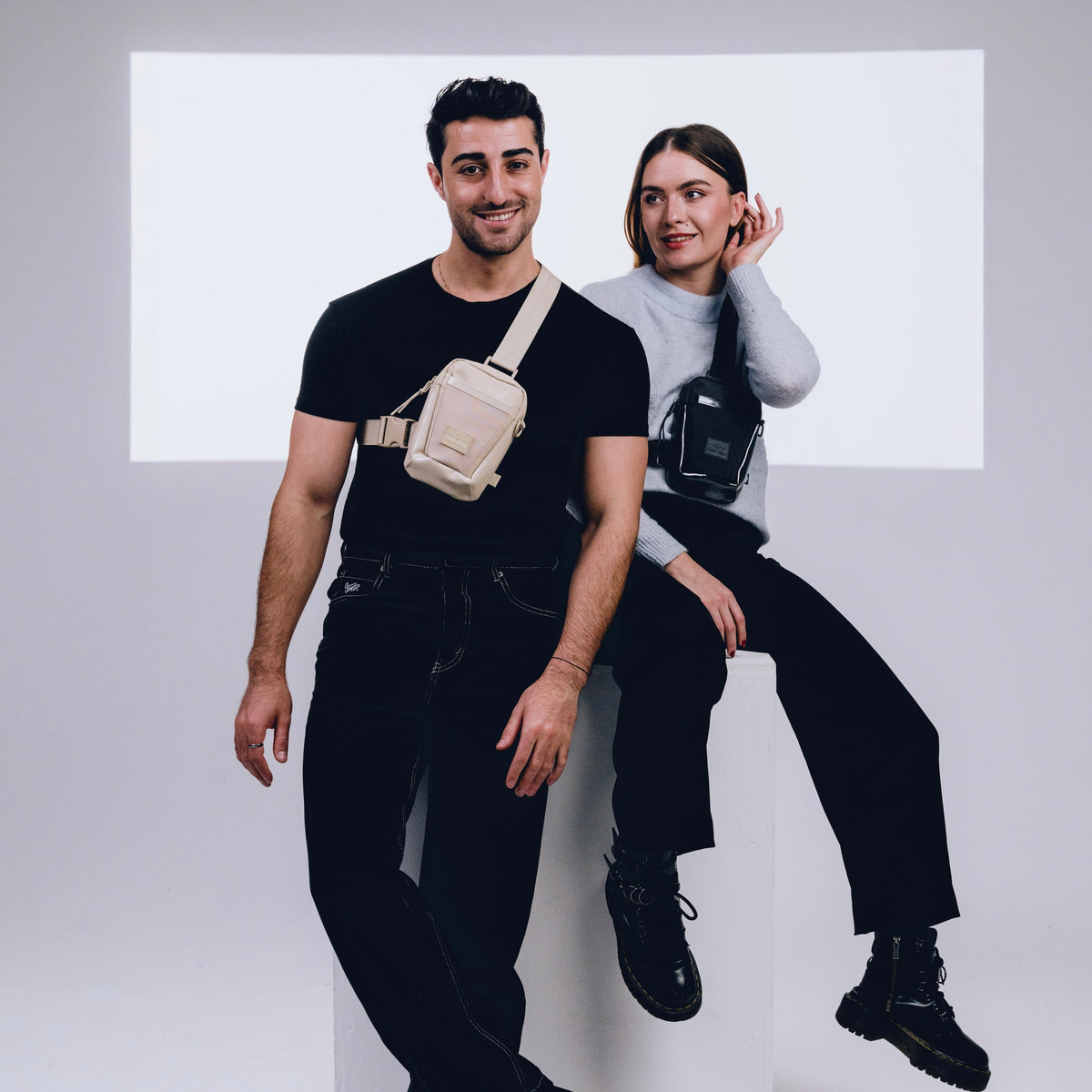 Mann mit schwarzem T-Shirt trägt beige Umhängetasche 'Lex', Frau mit grauem Pullover sitzt mit schwarzer 'Lex' Umhängetasche