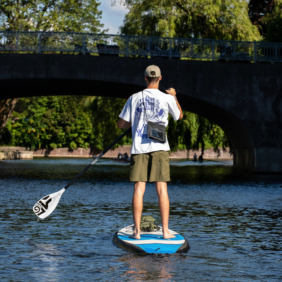 Rückansicht eines Mannes auf einem Stand-up-Paddle-Board mit Dry Bag 'Alfie' am Körper, umgeben von Wasser und Brücke im Hintergrund
