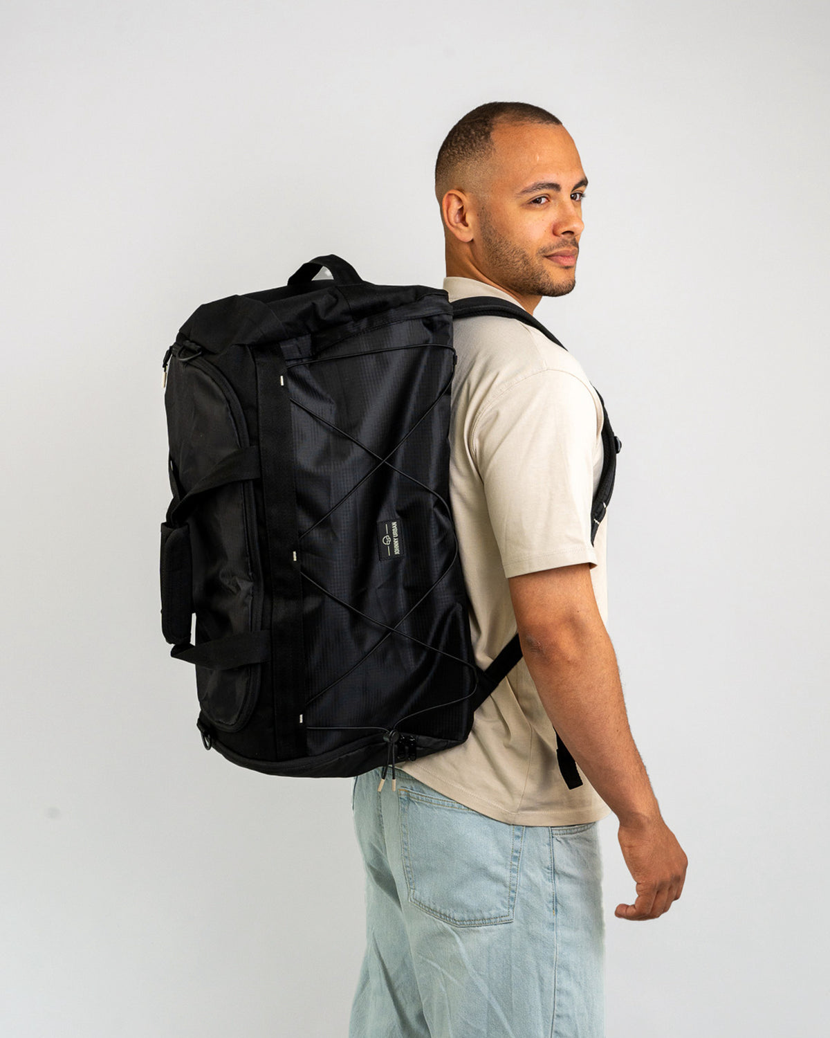 Mann trägt schwarzen Duffle Bag 'Shawn Large' auf Rücken, beige T-Shirt, helle Jeans