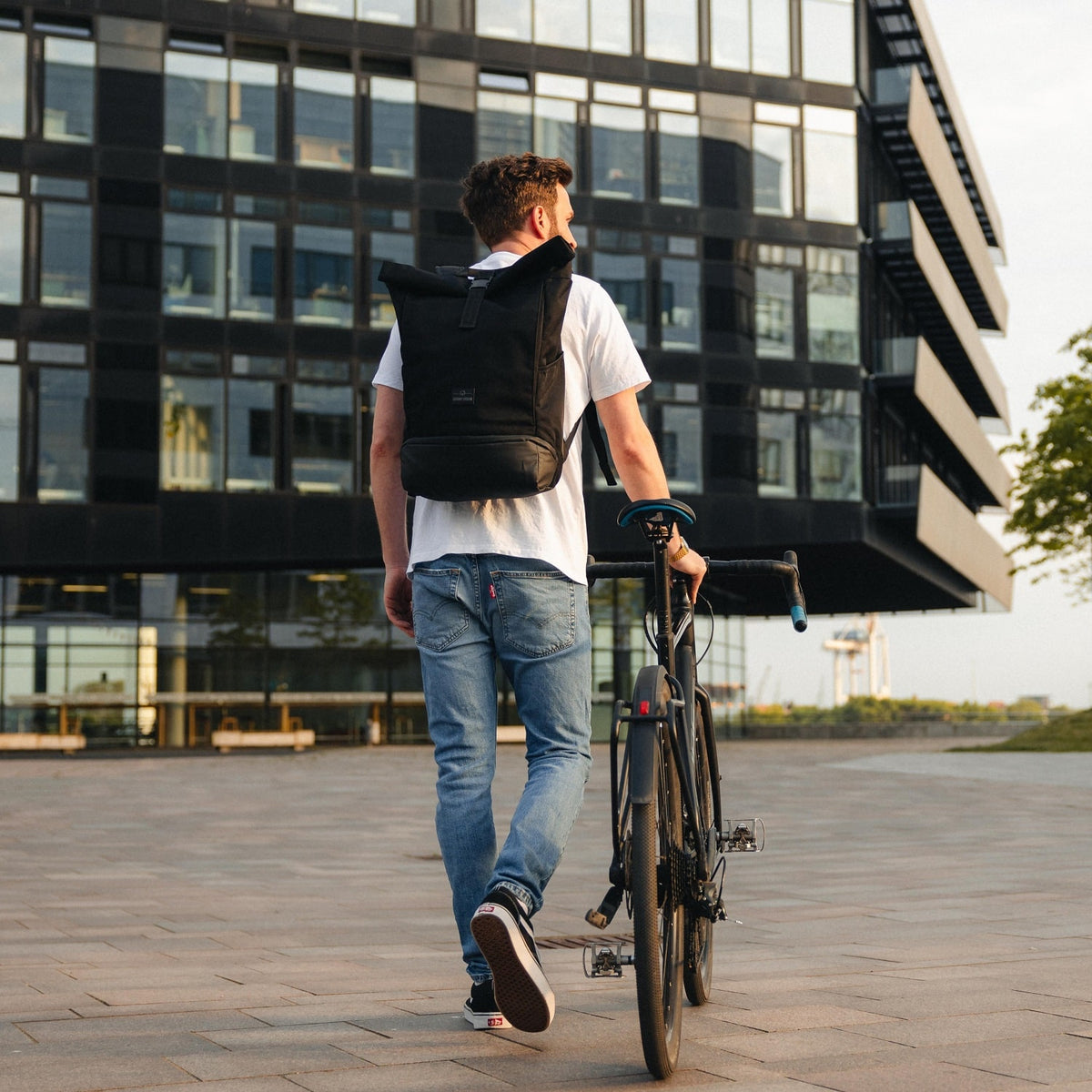 Mann trägt schwarzen Rolltop Rucksack ‚Allen Large‘ von Johnny Urban und schiebt Fahrrad vor modernem Gebäude