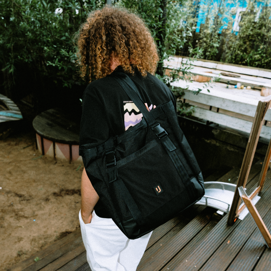 Person mit lockigem Haar trägt schwarze Johnny Urban Tote Bag 'Jessy' am Rücken an einem Outdoor-Festivalort