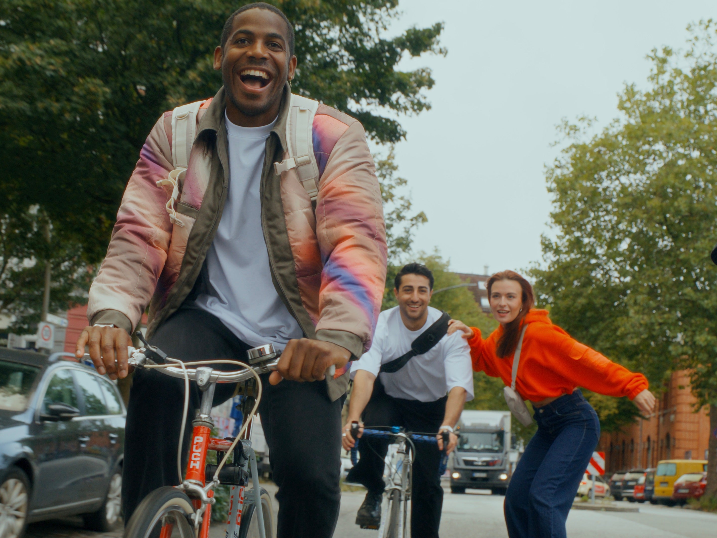 Zwei Männer auf Fahrrädern und eine Frau auf einem Skateboard fahren gemeinsam durch die Stadt und tragen Johnny-Urban-Klamotten.