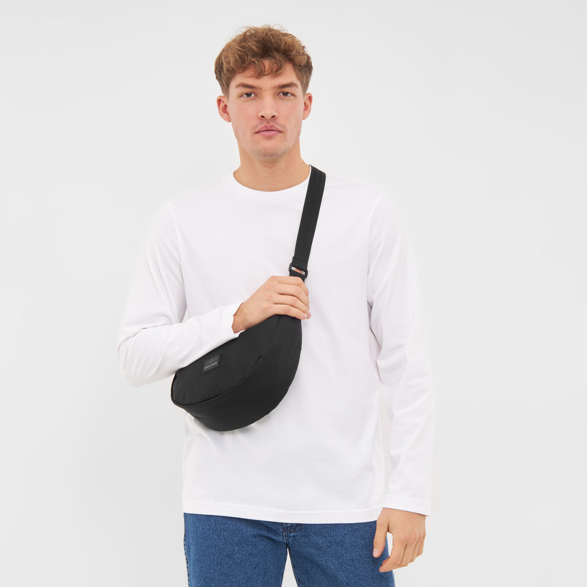 Mann mit weißem Langarmshirt und blauen Jeans trägt schwarze Half Moon Bag 'Ally Medium' quer am Oberkörper