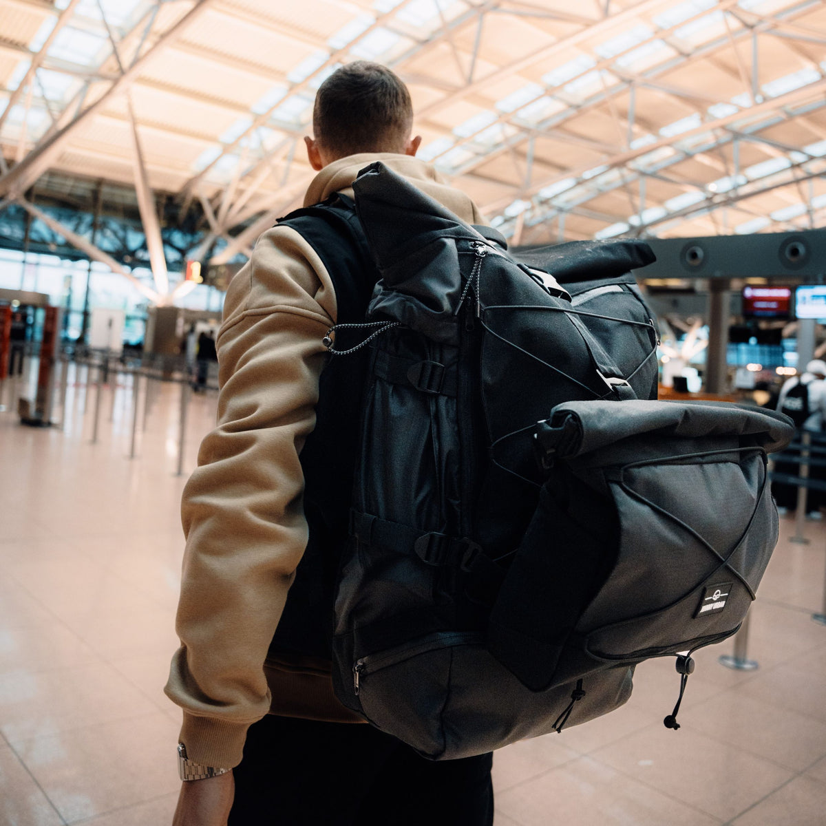 Mann mit beigefarbenem Pullover trägt schwarzen Reiserucksack 'Allen XL Travel' am Flughafen