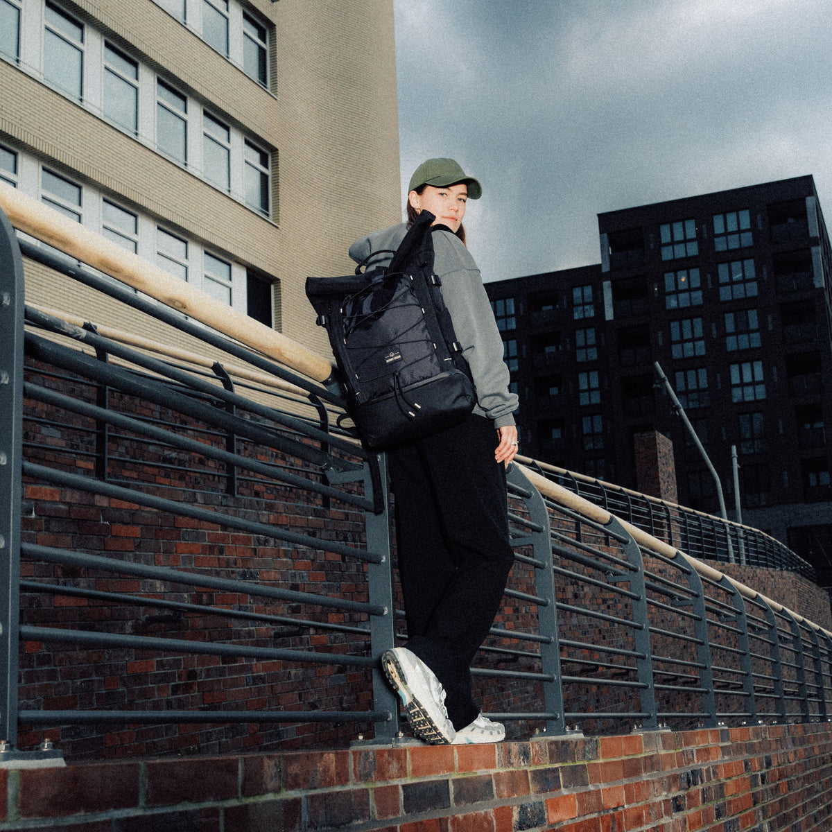 Frau mit grünem Cap trägt schwarzen Rolltop Rucksack 'Allen Large Move' urban vor Gebäuden