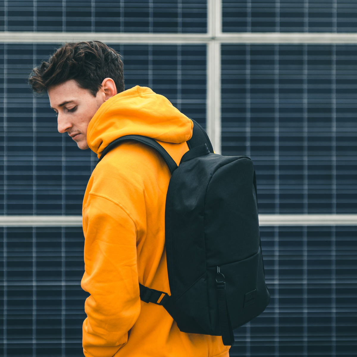 Junge trägt schwarzen Schulrucksack 'Milo' und orangenen Hoodie vor Solarzellenwand