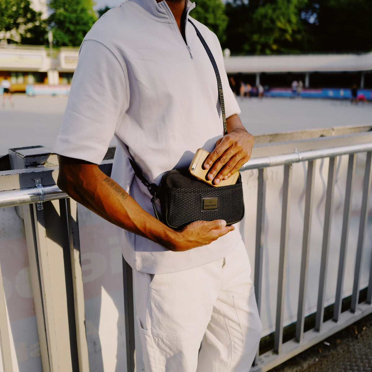 Person in hellbeiger Kleidung steckt ein Smartphone in die schwarze Umhängetasche 'Elliot' von Johnny Urban