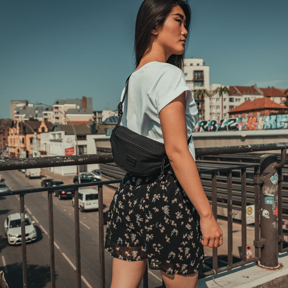 Frau mit schwarzer Bauchtasche 'Ben' von Johnny Urban, weißes T-Shirt, kurze schwarze Blumehose, städtische Hintergrund.