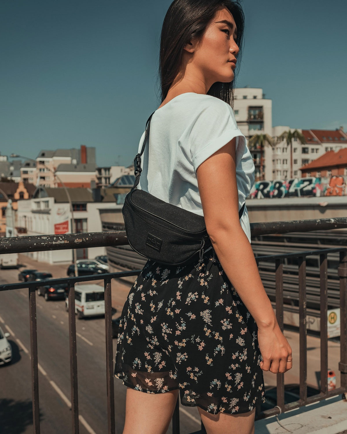 Frau mit schwarzer Bauchtasche 'Ben' von Johnny Urban, weißes T-Shirt, kurze schwarze Blumehose, städtische Hintergrund.