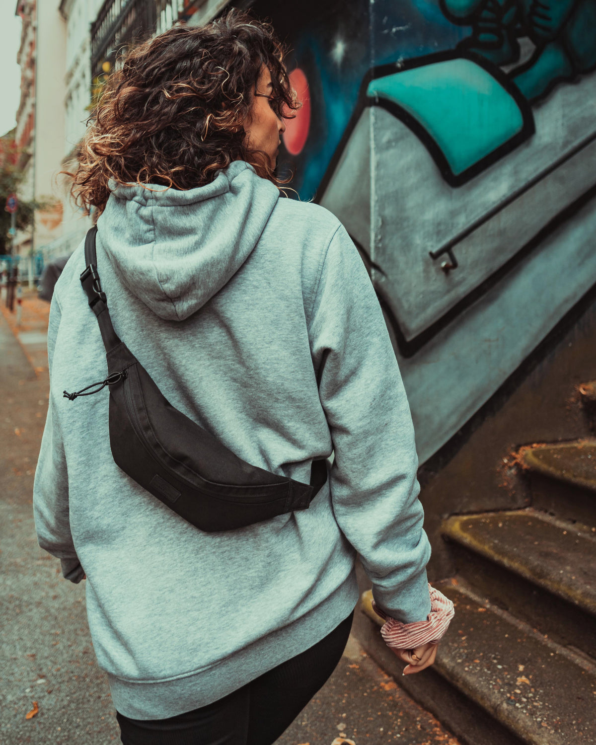 Frau mit lockigem Haar, grauem Hoodie und schwarzer Bauchtasche ‚Erik Small‘ trägt Tasche über Rücken in urbaner Umgebung