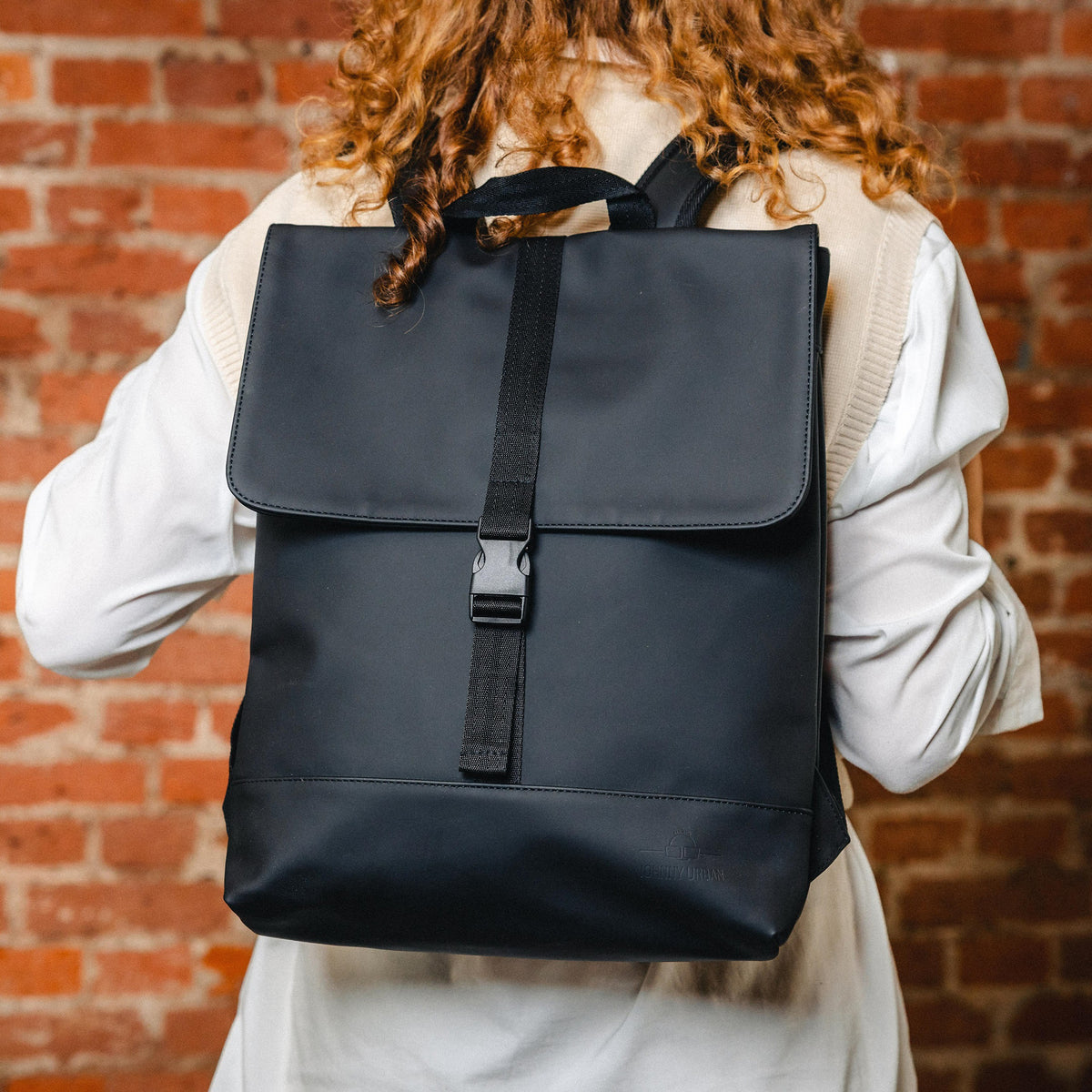 Schwarzer Damenrucksack ‚Ruby‘ von Johnny Urban an einer Person mit lockigem Haar vor roter Backsteinwand