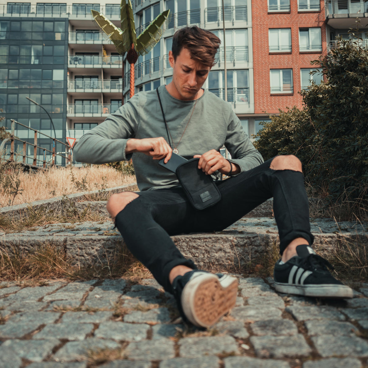 Junger Mann sitzt auf Steintreppe, trägt schwarze Umhängetasche 'Josh' und zieht etwas heraus