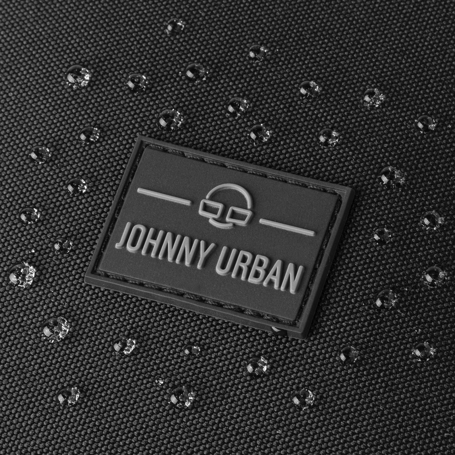 Nahaufnahme des Logos von Johnny Urban auf dem wasserfesten Stoff des Rucksacks ‚Nico‘ mit Wassertropfen