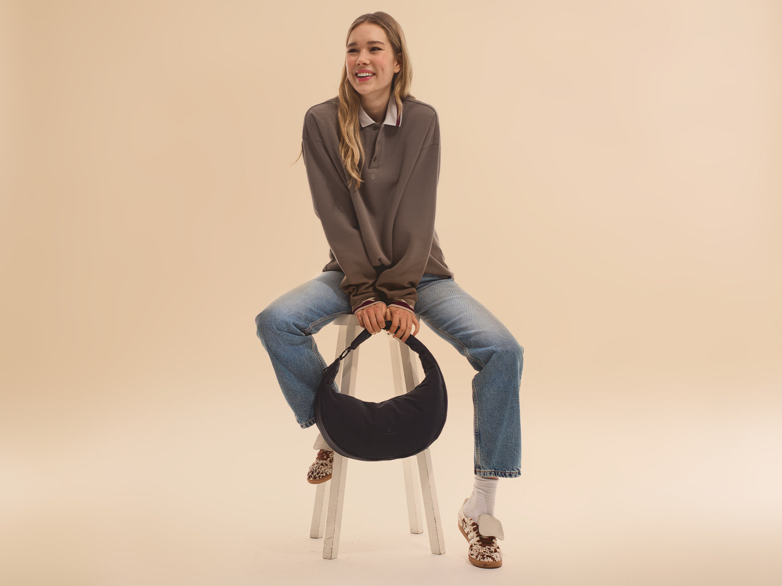 Frau sitze auf einem Hocker, lächelt  und hält eine schwarze Half Moon Bag "Lou Large" in den Händen.