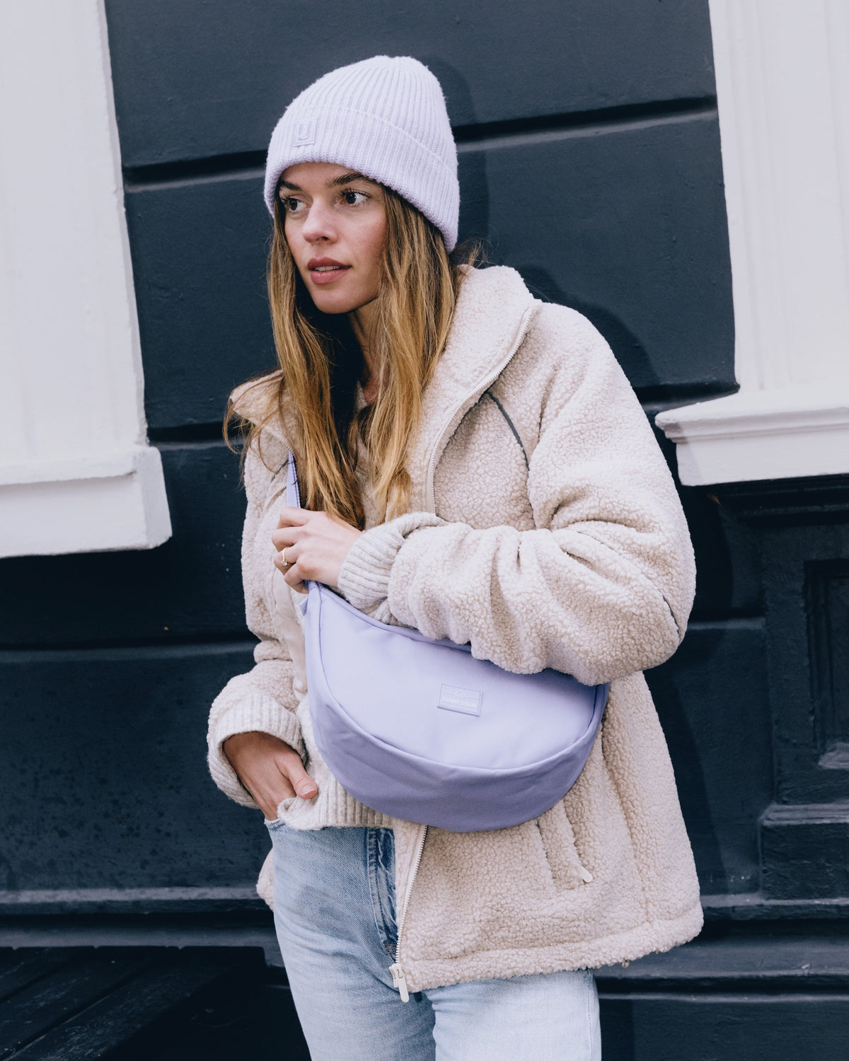 Frau mit hellviolettem Half Moon Bag 'Ally Medium', beiger Teddyjacke und Mütze vor schwarz-weißer Wand