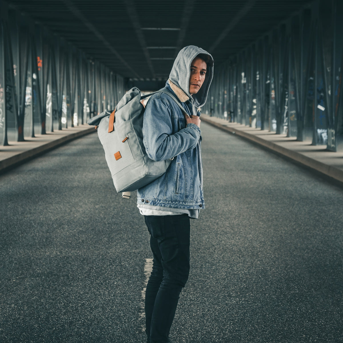 Junger Mann trägt grauen Rolltop Rucksack 'Adam Large' und Jeansjacke mit Kapuzenpullover unter einer Brücke