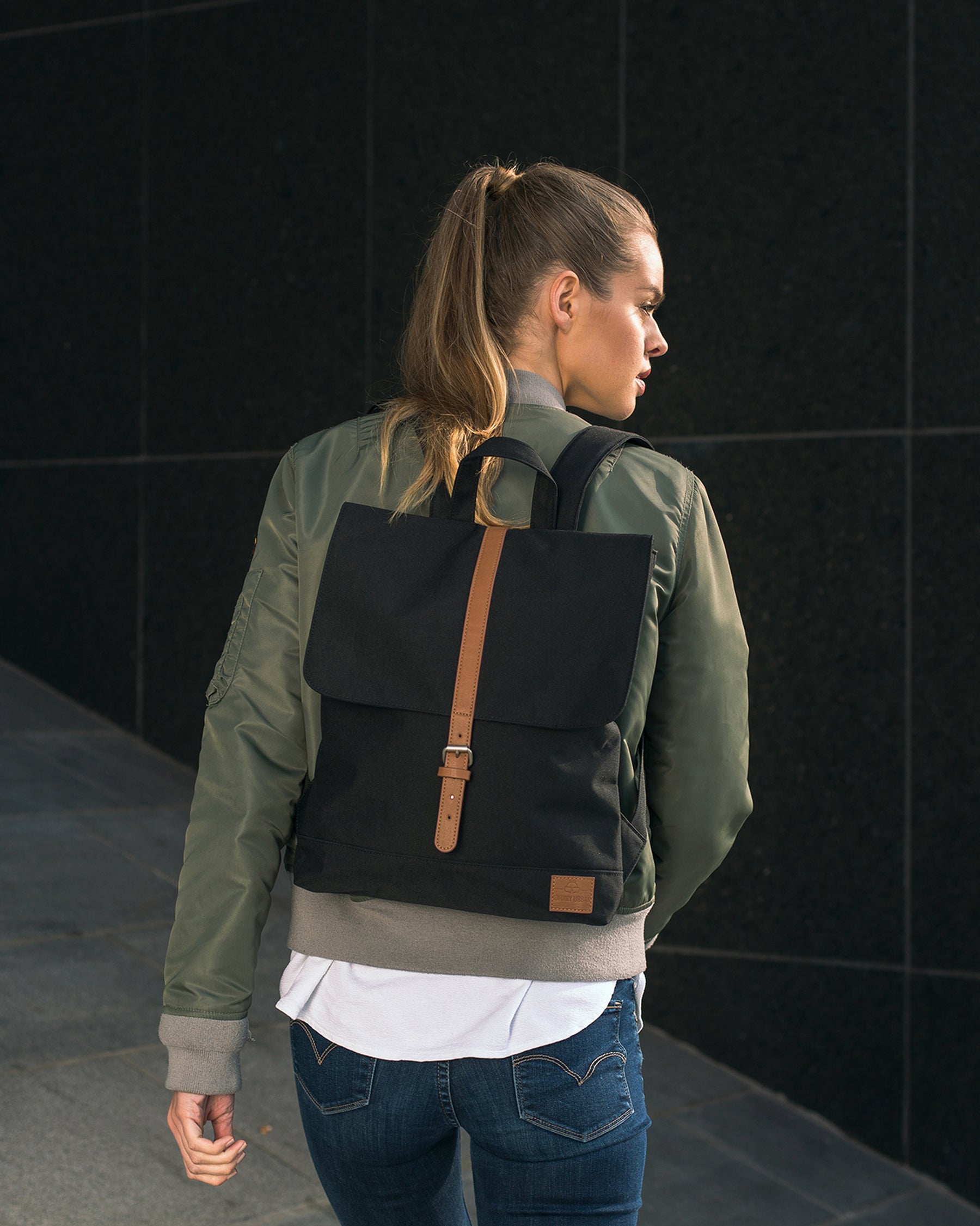 Frau trägt schwarzen Damenrucksack 'Mia' mit braunem Lederriemen in urbaner Umgebung