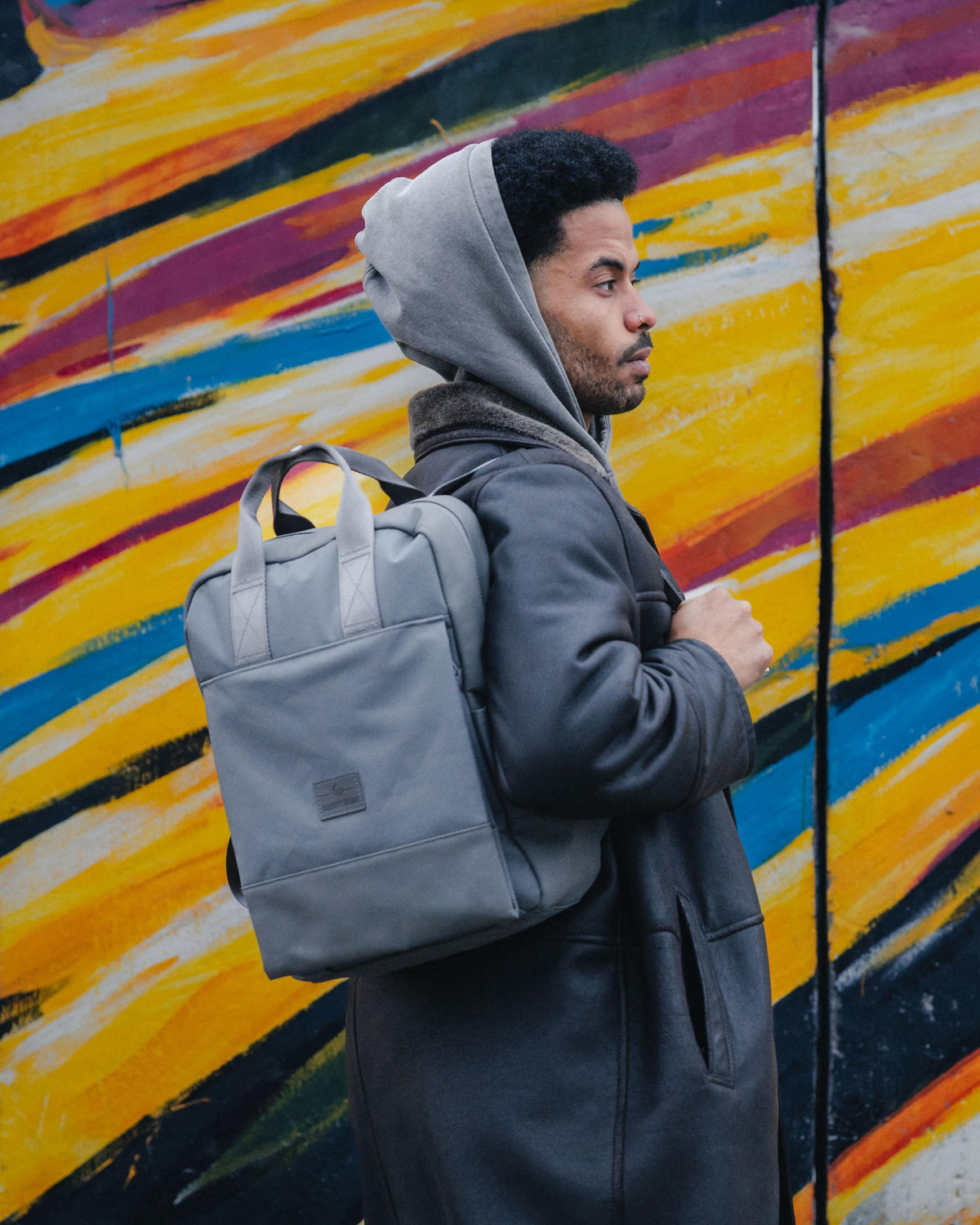 Mann mit grauem Rucksack 'Jona Large' steht vor einer bunten Wand mit gelben, blauen und roten Streifen
